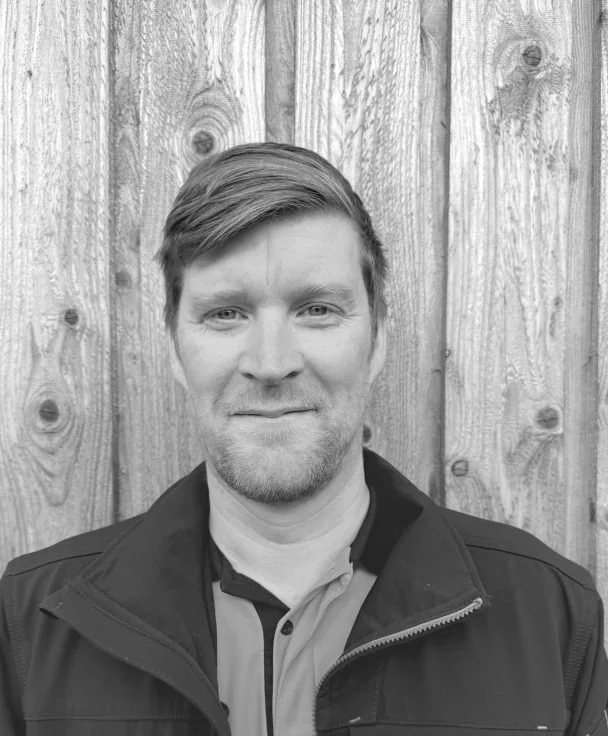 Black and white photo of a smiling man with short hair and a beard, wearing a collared shirt and a zip-up jacket, standing in front of a wooden fence.