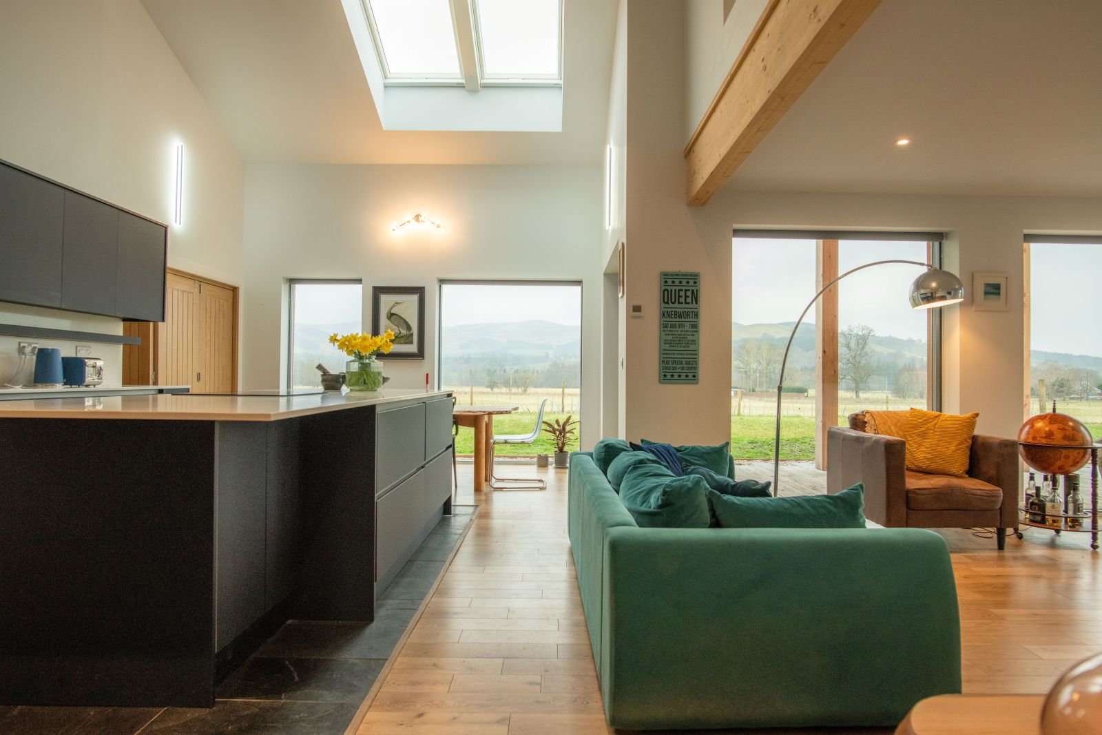 MAKAR home living room with a green couch, armchair, floor lamp, large windows showing a view of countryside, skylight, and modern decor.