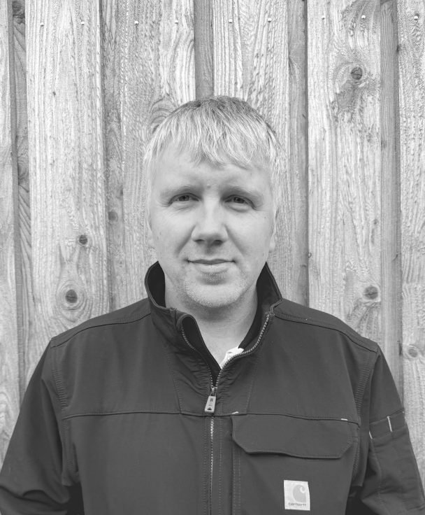 A man with short blond hair wearing a black jacket, standing in front of a wooden fence.