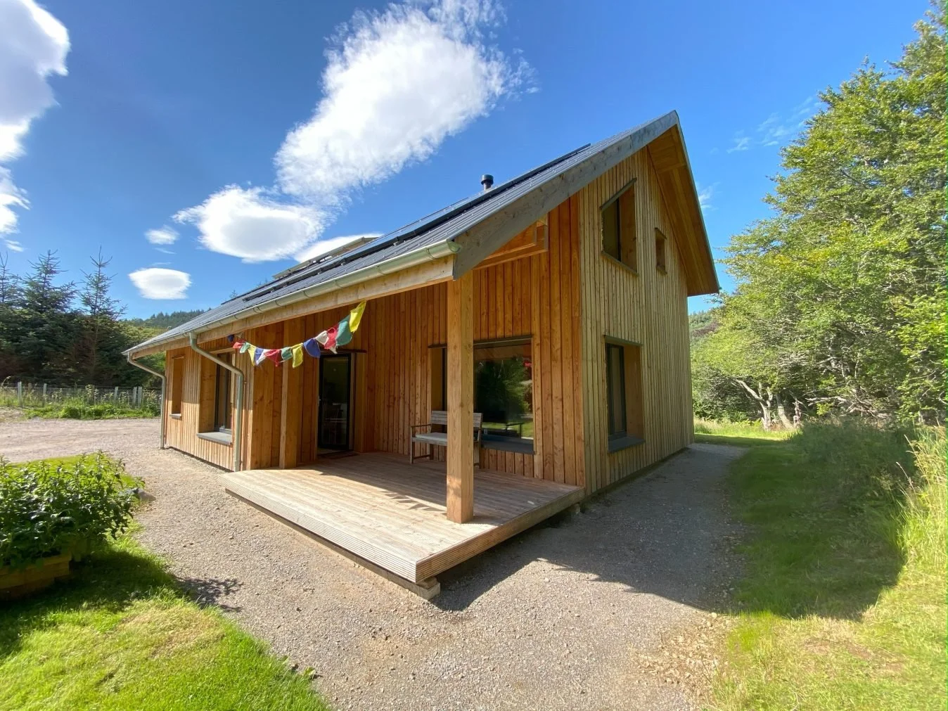 A MAKAR Timber home with a small deck in a rural setting, surrounded by green grass and trees, under a bright blue sky with clouds.