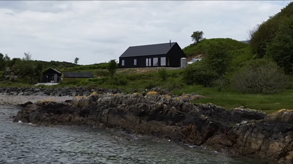 A coastal scene on Skye with a black MAKAR home on a grassy hill, rocks along the shoreline, and the sea in the foreground.