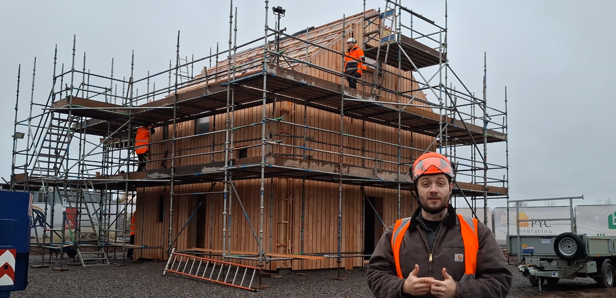 MAKAR construction site with workers building a wooden house, scaffolding surrounds the house, and a man in a hard hat and safety vest is in the foreground about to talk to the camera.