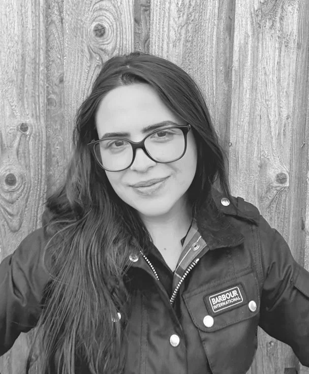 Young woman with long hair, glasses, and a jacket standing in front of a wooden fence.