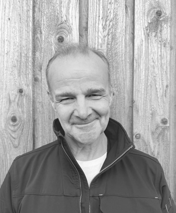 A man with light skin, short hair, and prominent facial features, smiling with closed eyes, is standing in front of a wooden fence. He is wearing a dark jacket over a white shirt.