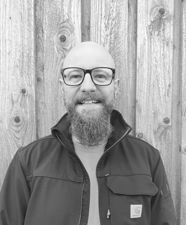 A man with glasses, a bald head, a beard, and a mustache, smiling in front of a wooden fence.