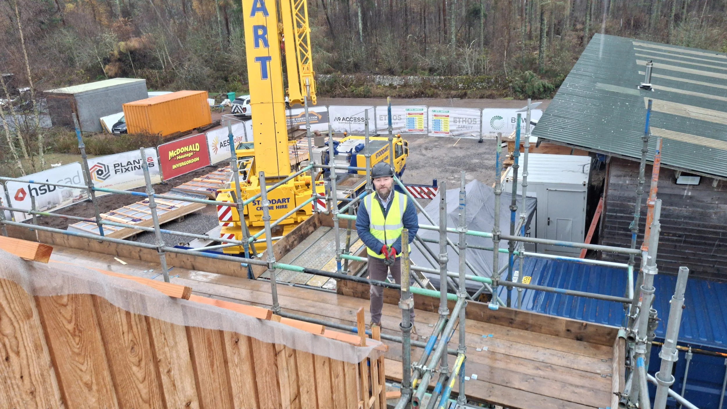 A construction worker wearing a safety vest and helmet stands on scaffolding at a building site, with a large yellow crane and construction materials in the background.