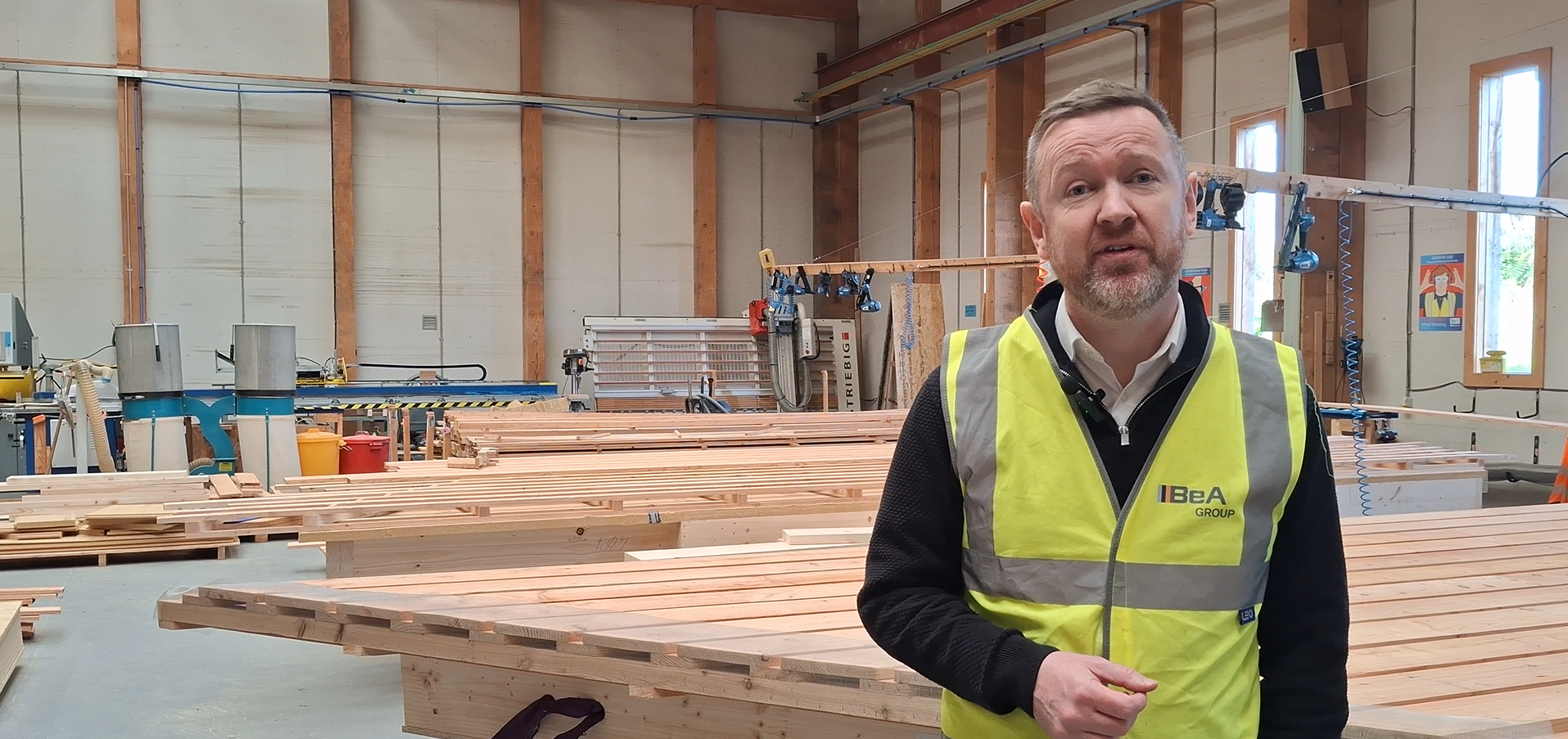 A man wearing a yellow high-visibility safety vest standing inside a workshop with stacks of wooden planks and woodworking equipment in the background.