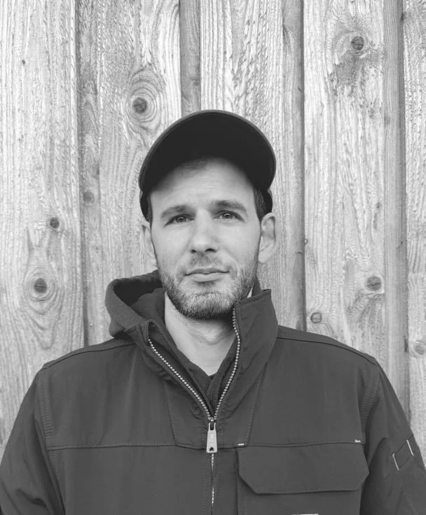 A man with light skin, short hair, and facial hair standing outdoors in front of a wooden fence, wearing a cap and a dark zip-up jacket.