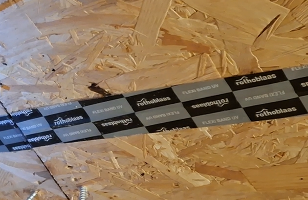 Close-up of a black and white roofing tape labeled 'rothoblaas' and 'FLEXI BAND UV' attached to wooden OSB sheathing.