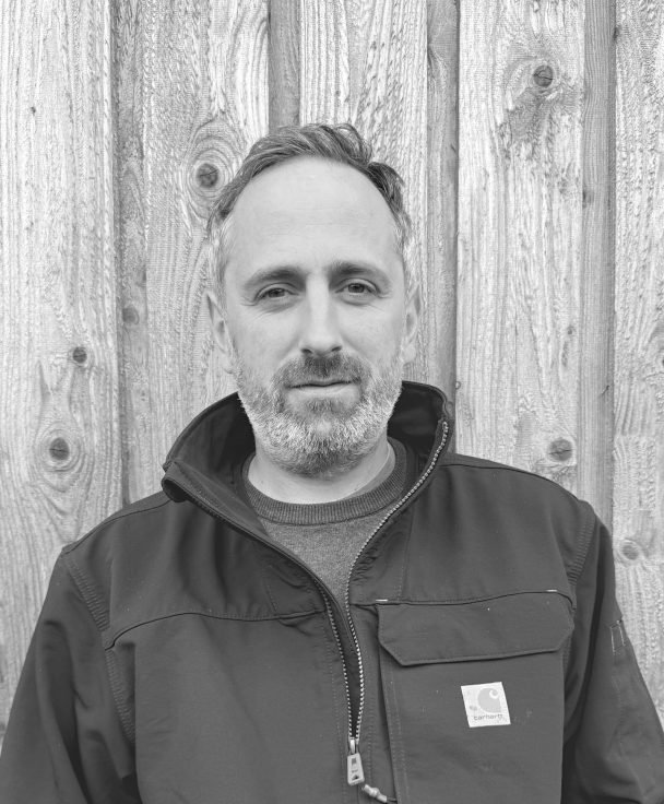 A man with short hair and a beard standing in front of a wooden fence, wearing a dark jacket.