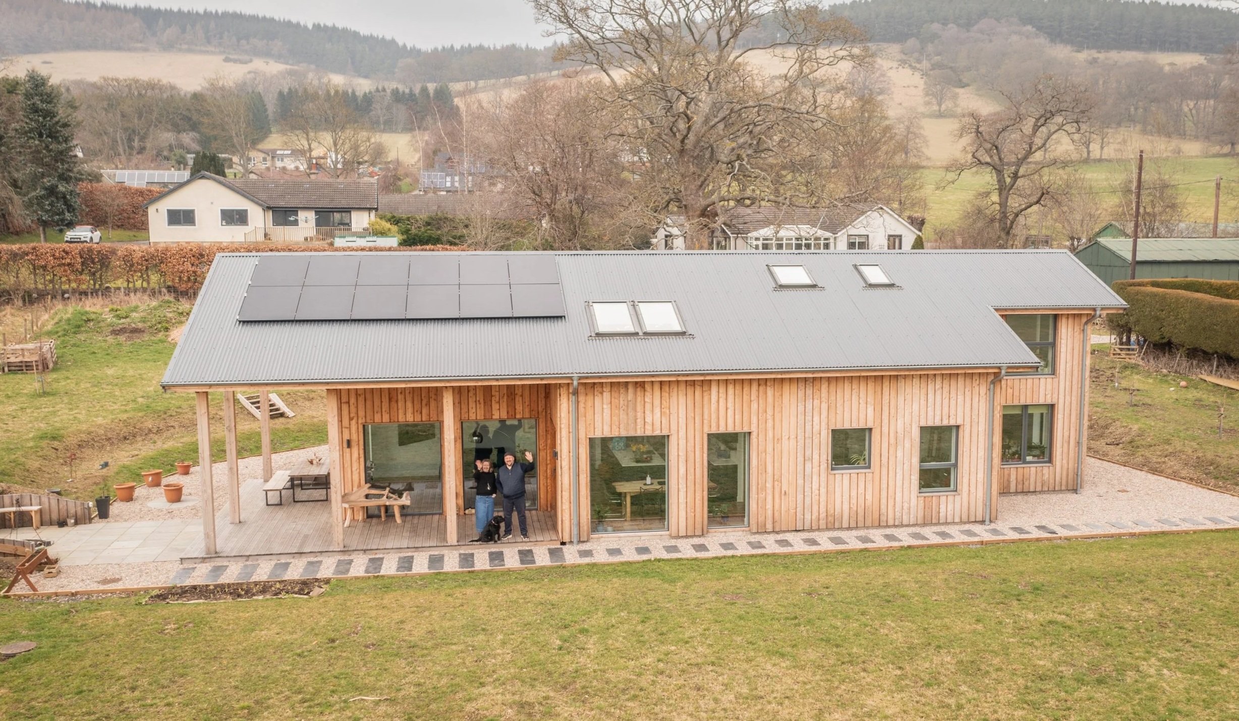 A modern wooden house with a metal roof, large front windows, and two people waving from the porch on a cloudy day.