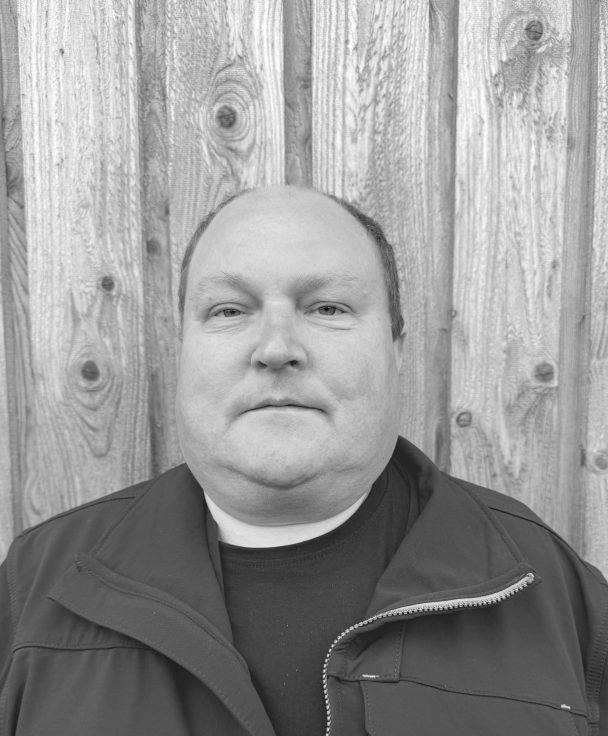 Black and white photo of a man with a serious expression, standing in front of a wooden fence.