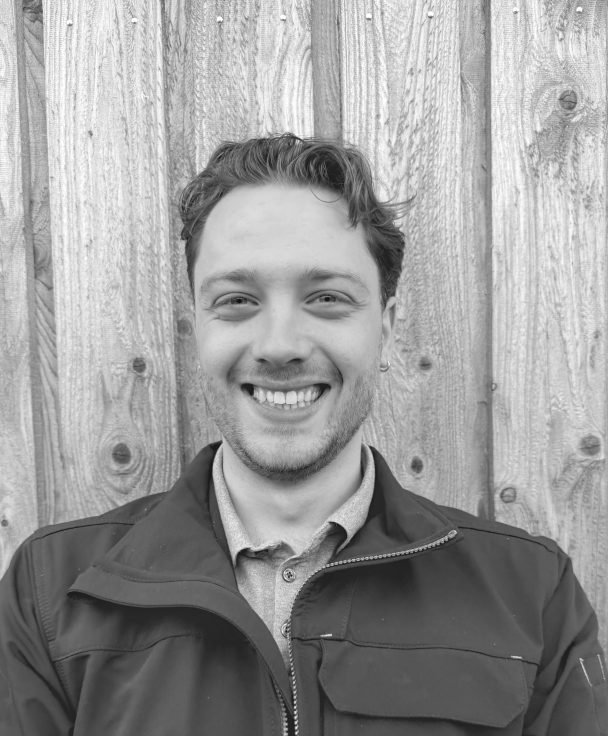 A smiling man with short wavy hair wearing a zip-up jacket over a collared shirt, standing in front of a wooden fence.