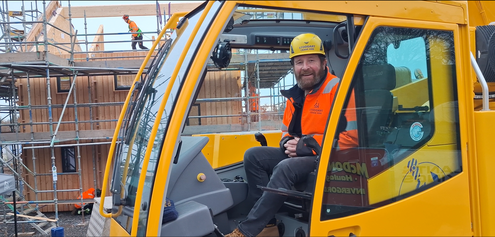 A smiling construction worker with a beard, wearing a yellow safety helmet and an orange safety vest, sitting inside a yellow construction vehicle at a building site framed with scaffolding and wood structures.
