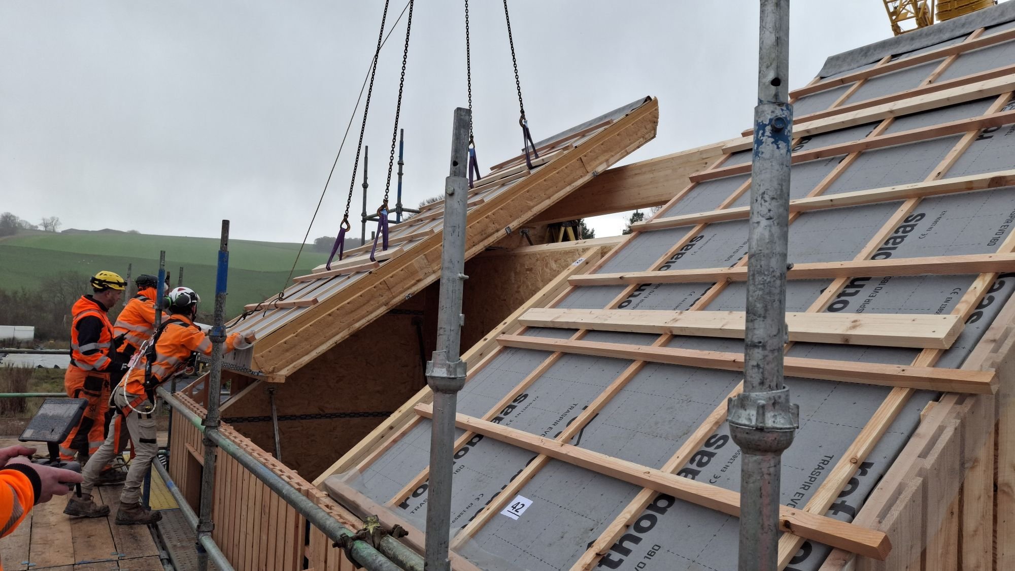 MAKAR Construction workers installing timber roof panels with natural insulation, which have been constructed off-site, onto a sloped roof with wooden framing, scaffolding, and a crane in a rural area.