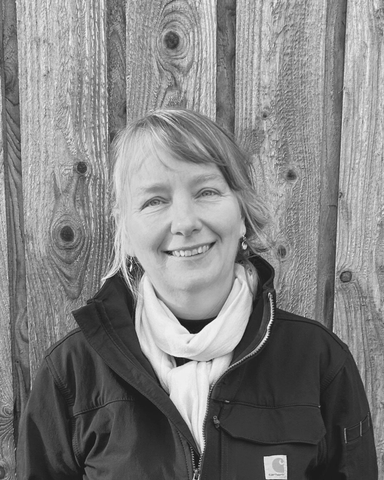 A woman with light-colored hair smiling, wearing a dark jacket and a light scarf, standing in front of a wooden fence.