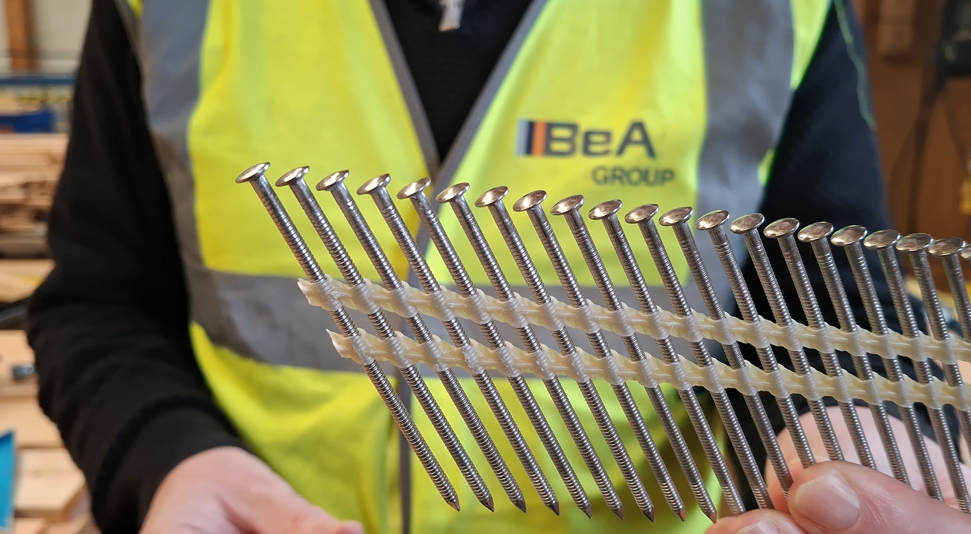Person holding a branded nail sample in a workshop, wearing a yellow safety vest with 'BeA Group' logo