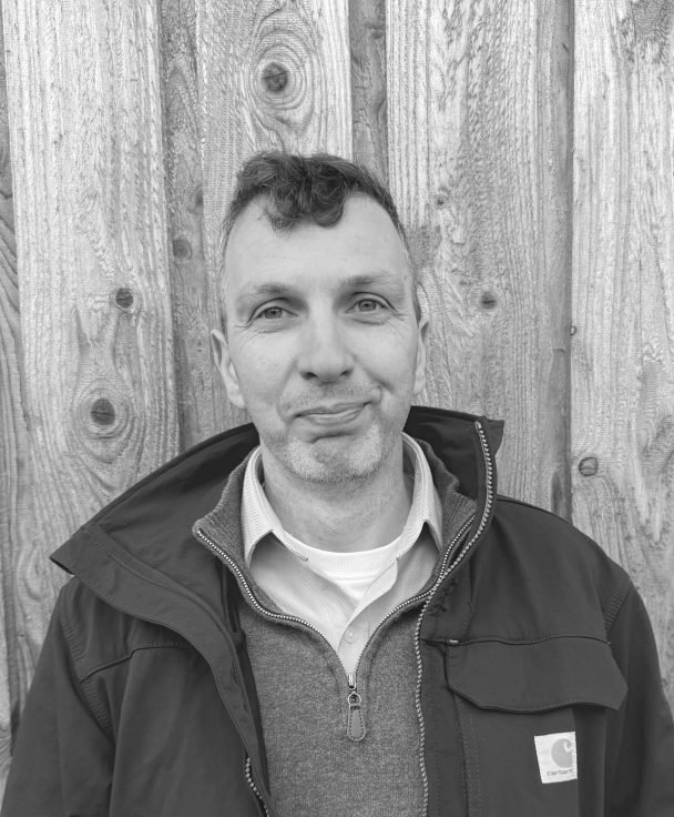 A man with short, curly hair, wearing a collared shirt, layered with a zippered sweater and a black Carhartt jacket, standing in front of a wooden fence.