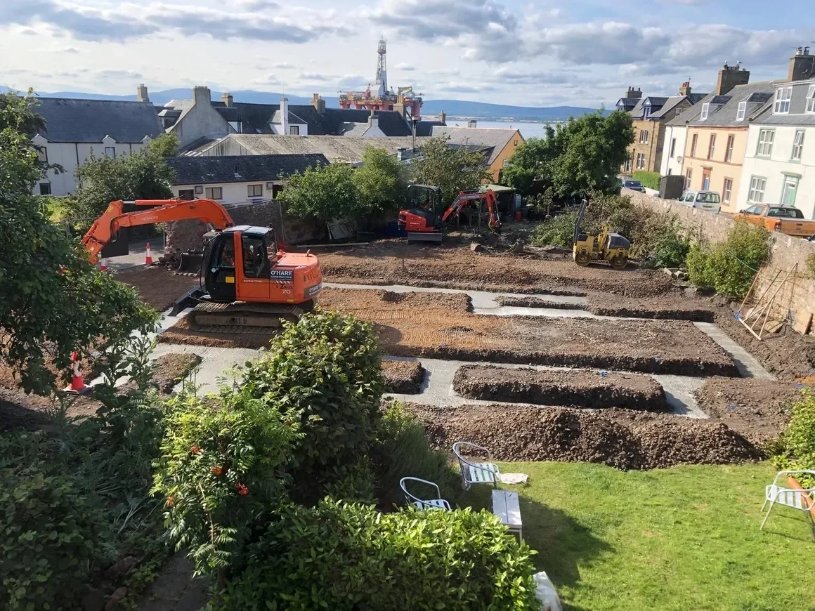A construction site in a residential area with excavators digging and leveling the ground. Visible in the background are houses, trees, and a body of water with some industrial structures such as a crane and a platform. There are chairs and a small table on a grassy area in the foreground.