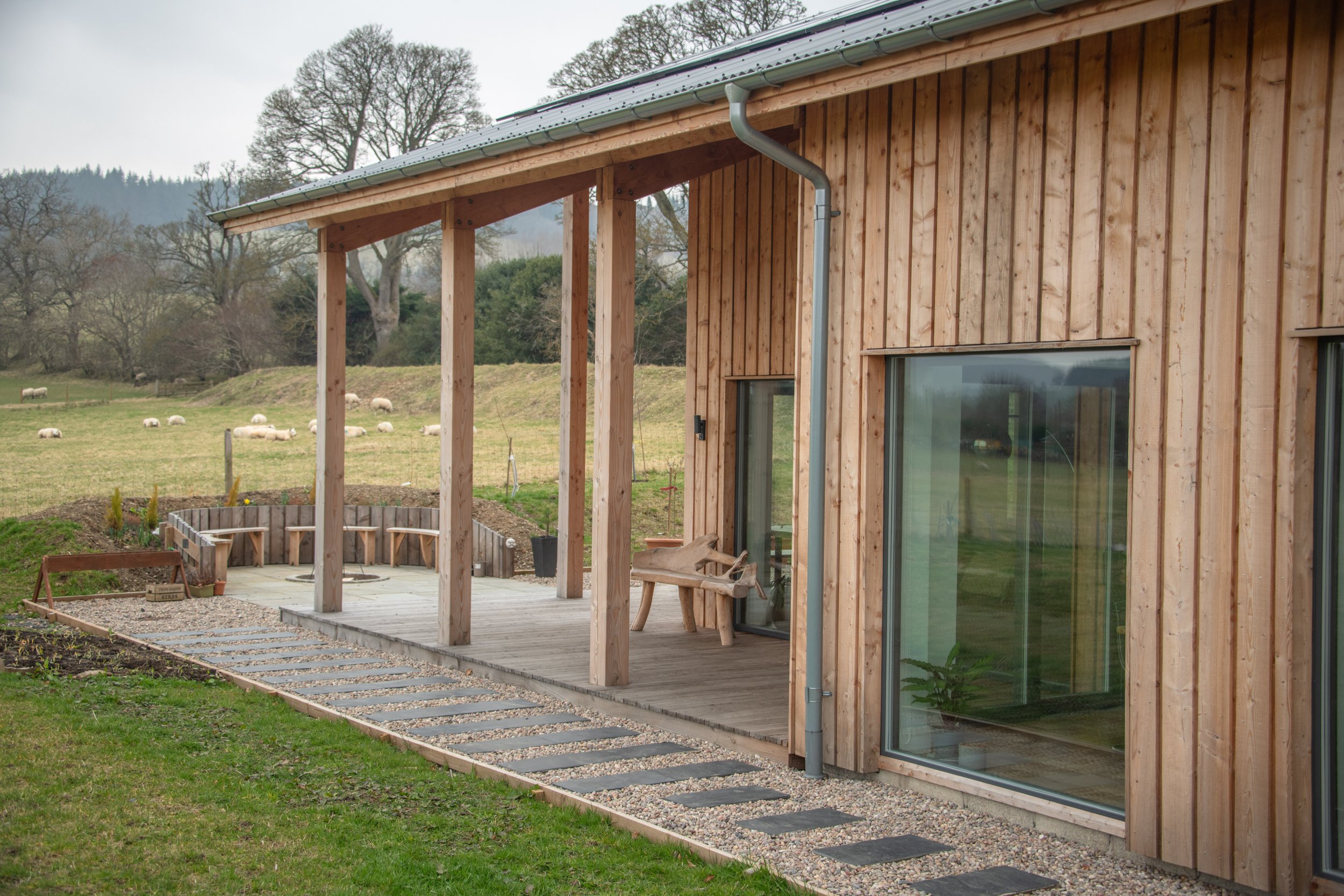 Wooden house with a small deck and large glass sliding door, overlooking a grassy field with sheep.