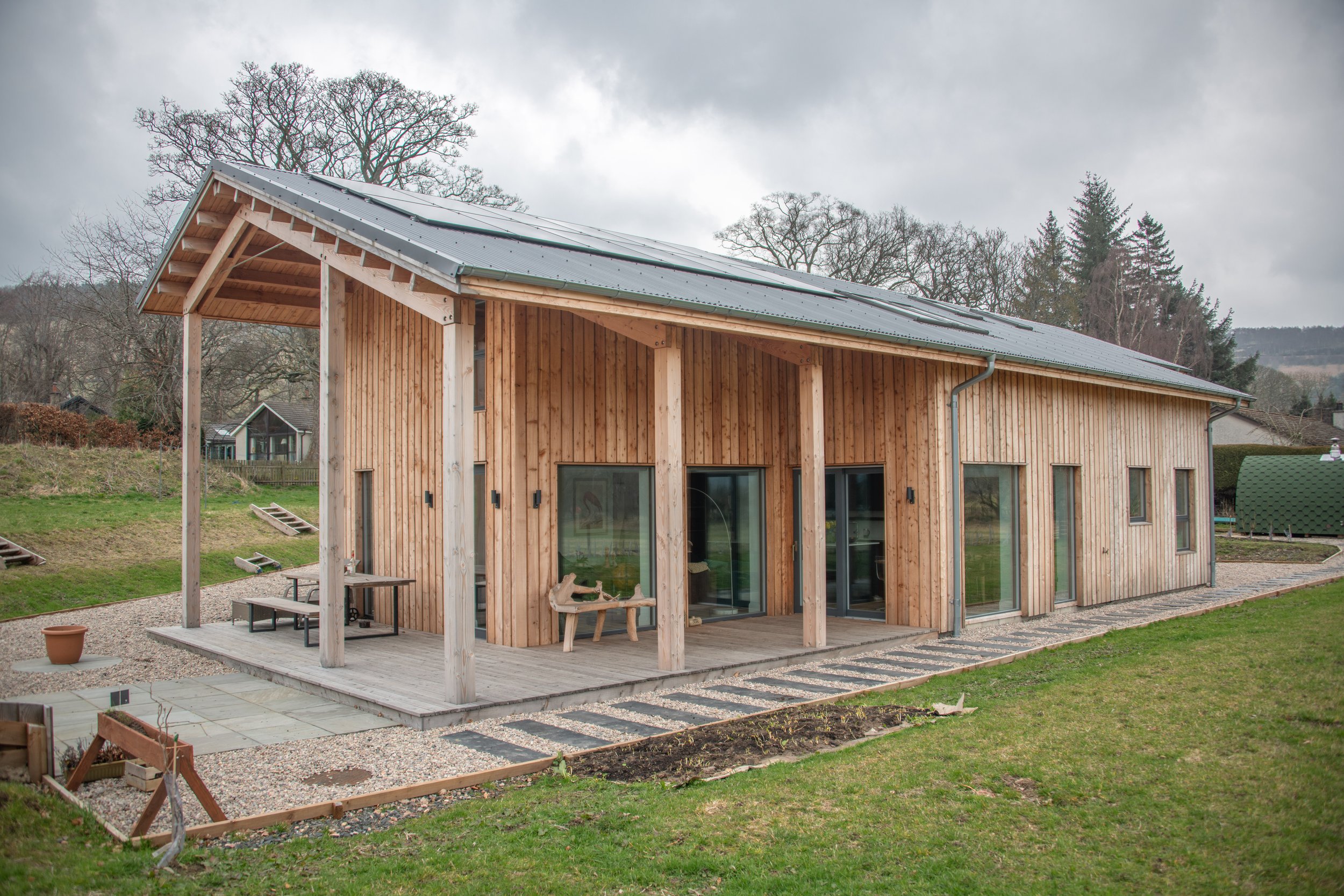 A modern timber MAKAR home with a metal roof, situated outdoors on a grassy area. The house has large windows and a small patio with benches and decor, surrounded by a landscaped yard with gravel and stepping stones.