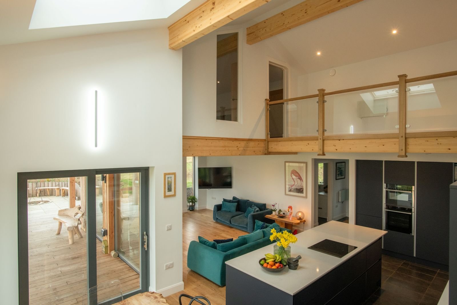 An open-concept living room with a kitchen, featuring a balcony with outdoor furniture, a blue couch, a TV, and artwork on the walls, with wooden accents and a high ceiling.