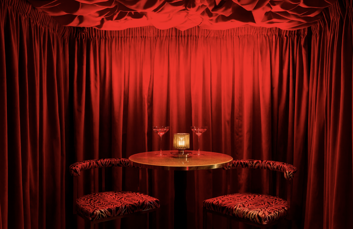 A small round table with two martini glasses and a candle, set in front of red curtains and upholstered chairs with a red and black patterned fabric.