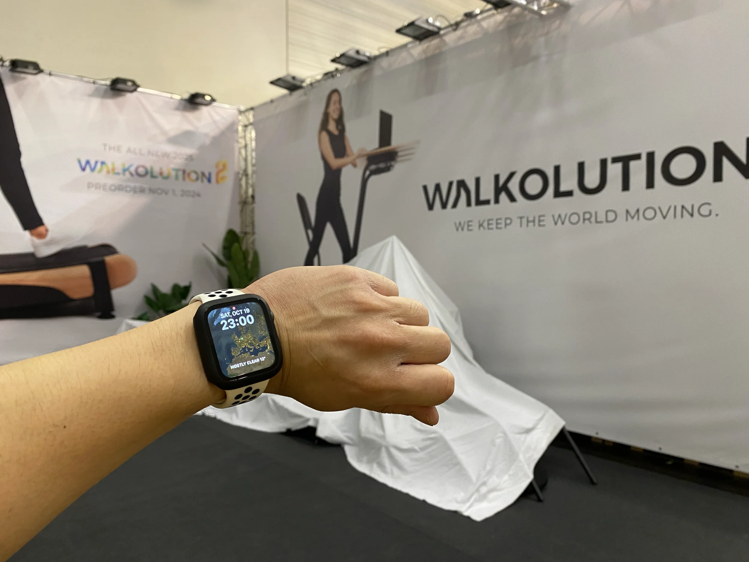 Close up of a wrist wearing a smartwatch showing late night time during Walkolution trade show setup, with branded booth walls, covered equipment, and lighting rigs in the background, capturing behind the scenes build progress.