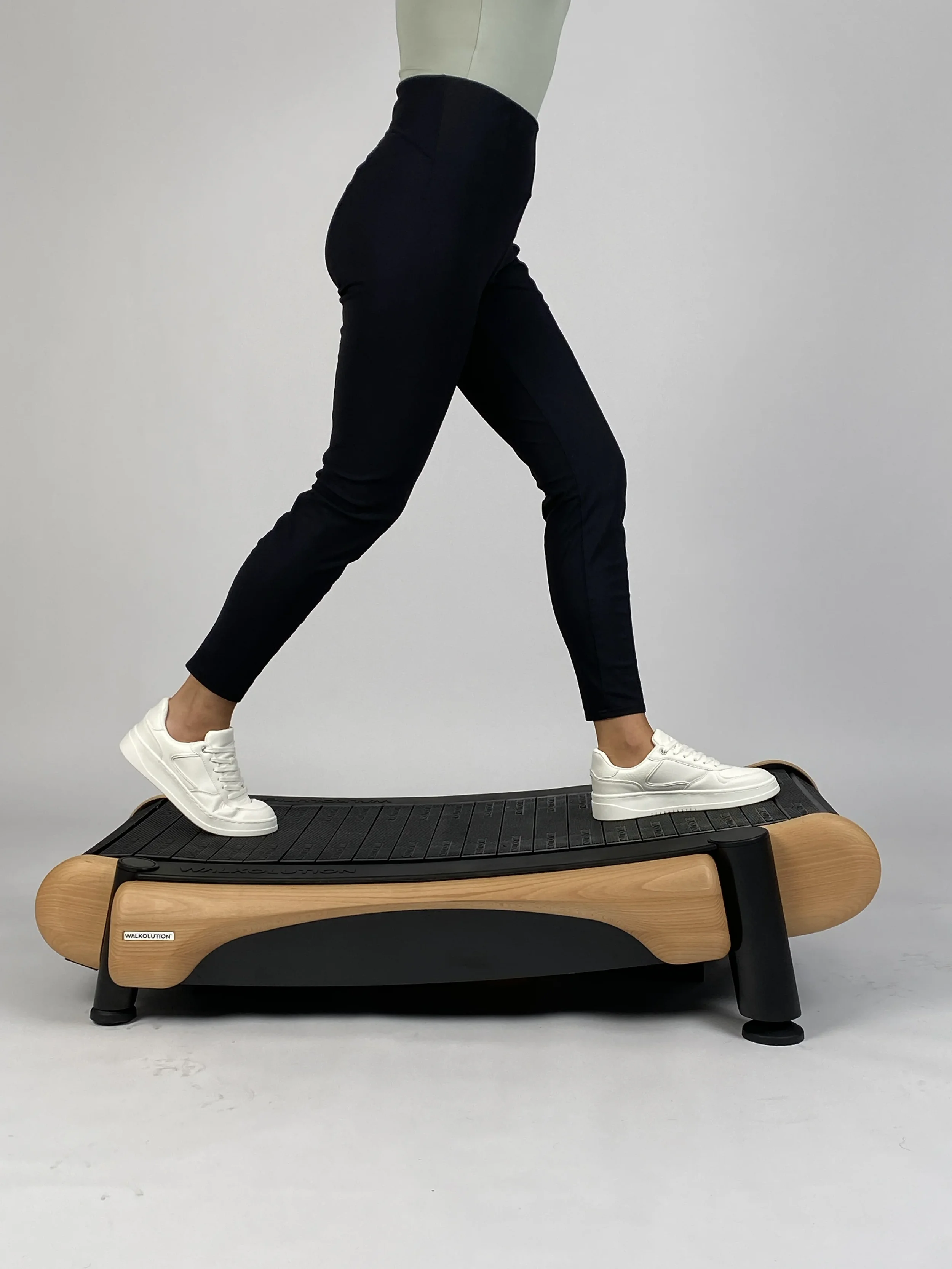 Person walking on a wooden manual treadmill desk wearing black pants and white sneakers against a clean studio background, highlighting smooth walking motion, ergonomic design, and active work movement.