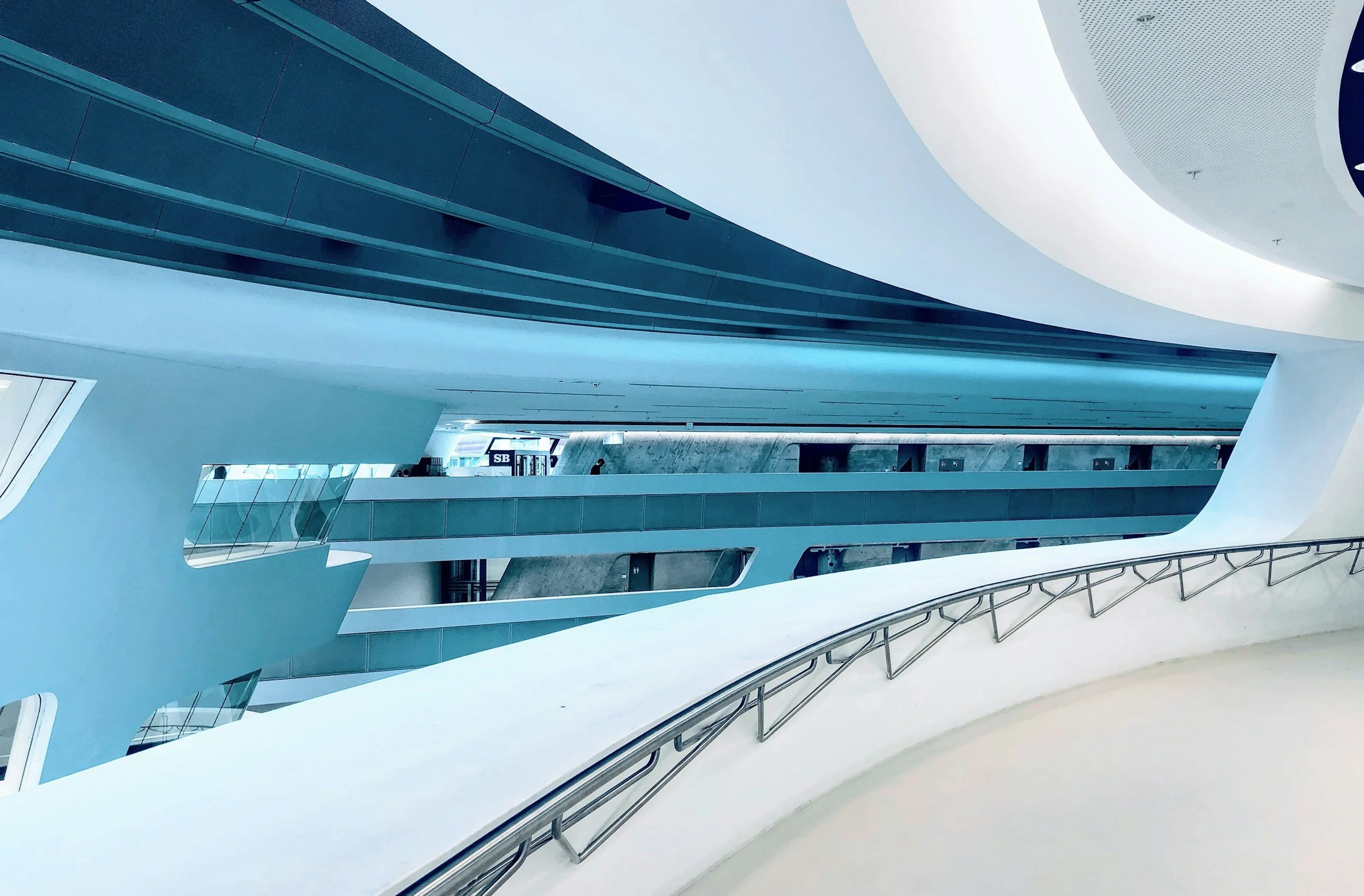 Intérieur moderne avec plusieurs niveaux, rampes, structures blanches et bleues, et des fenêtres en verre.