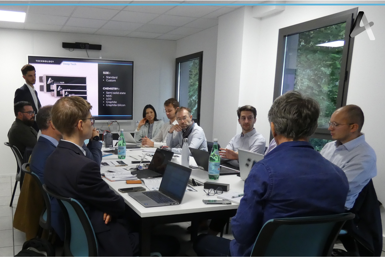 Une réunion de travail avec plusieurs personnes assises autour d'une table, une présentation sur un écran derrière elles et des fenêtres donnant sur des arbres