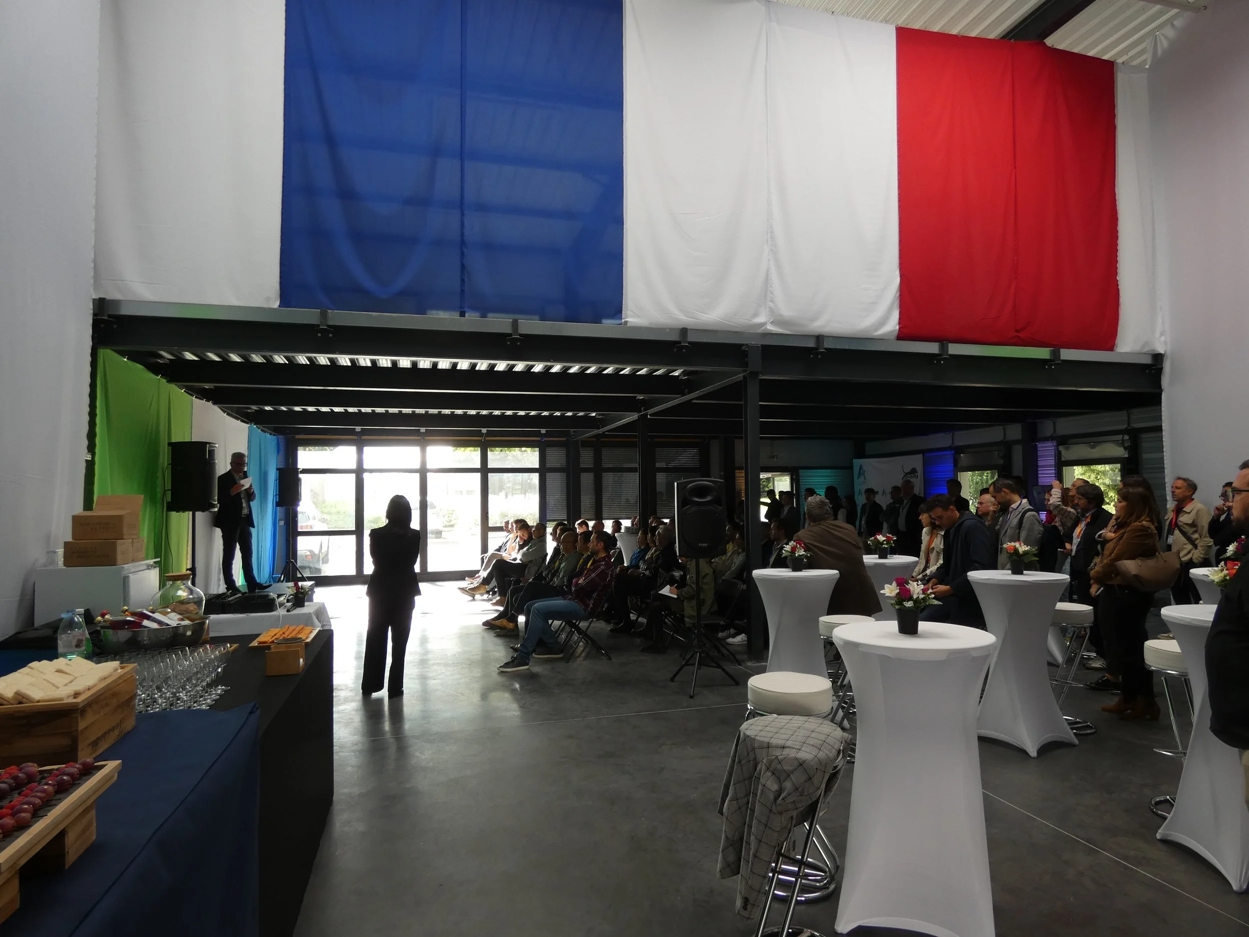Salle de réunion ou événement avec des gens assis et debout, décorée avec un drapeau tricolore français en haut, tables hautes avec fleurs, et un stand avec snacks à gauche.