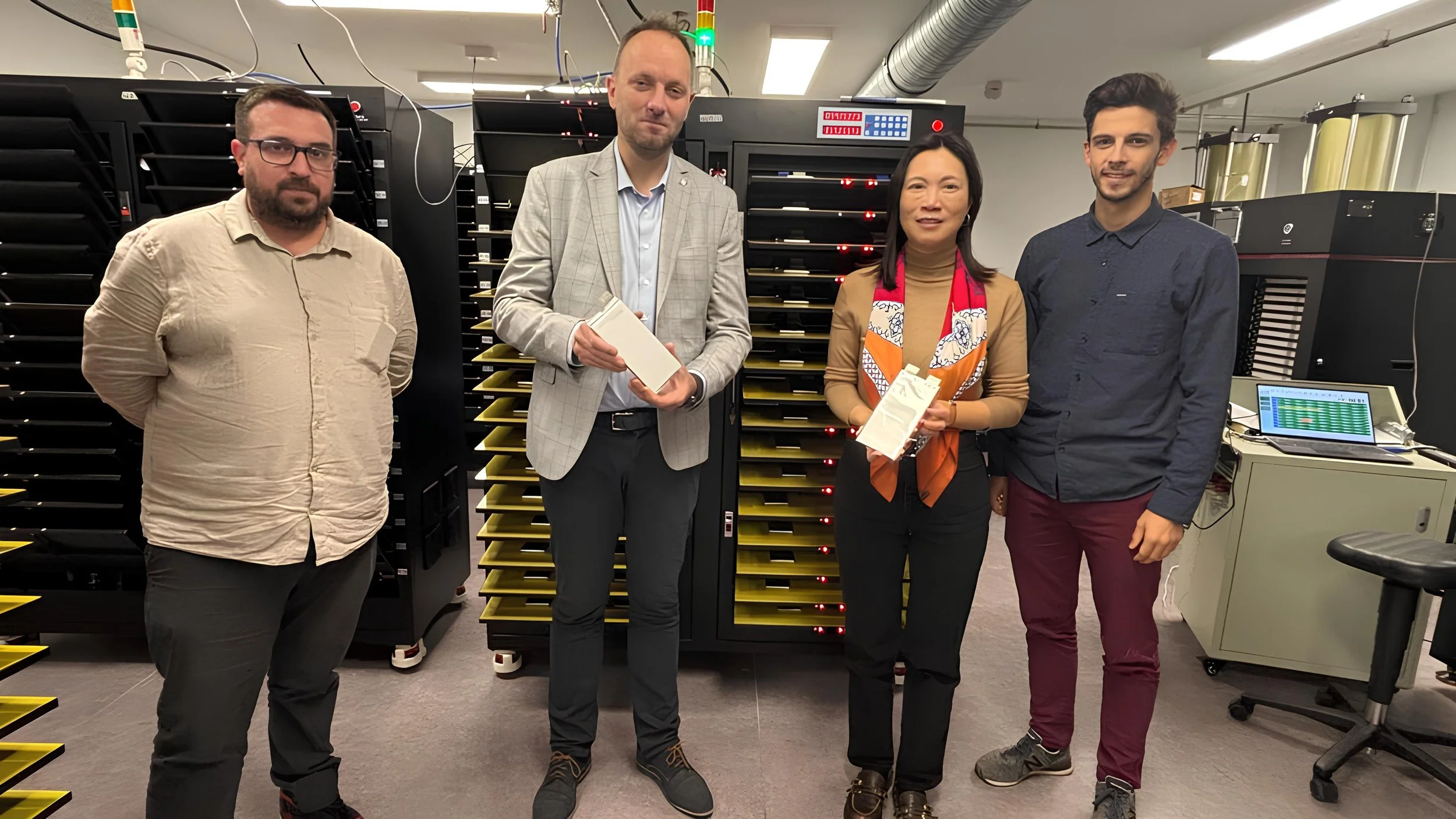 Cinq personnes debout dans une salle technique avec des serveurs en arrière-plan, deux hommes tenant des paquets.