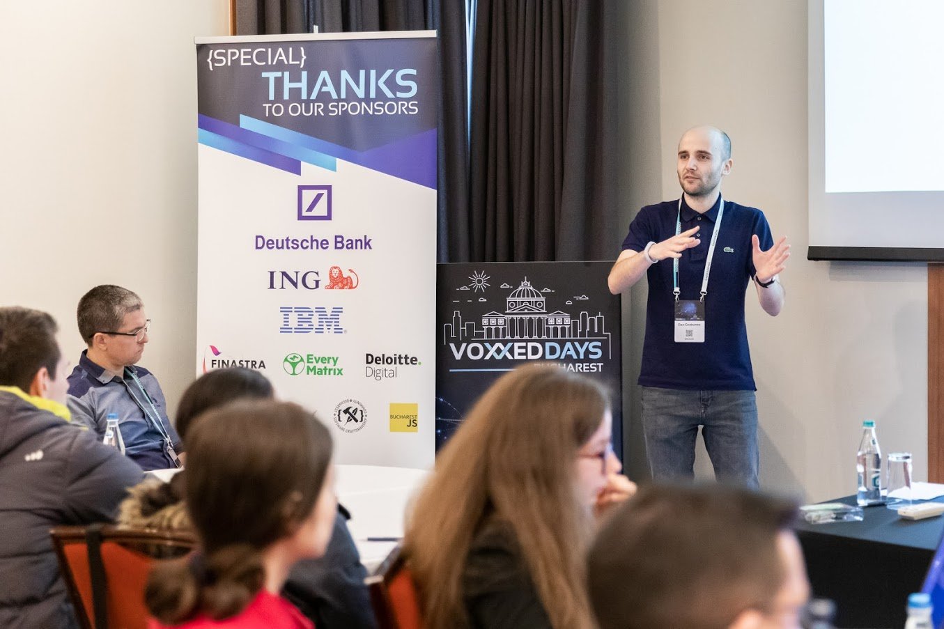 Dan Geabunea is standing and gesturing towards an audience at a conference, with a Voxxed Days Bucharest banner and a sponsor poster featuring Deutsche Bank, ING, IBM, and other logos visible behind him.