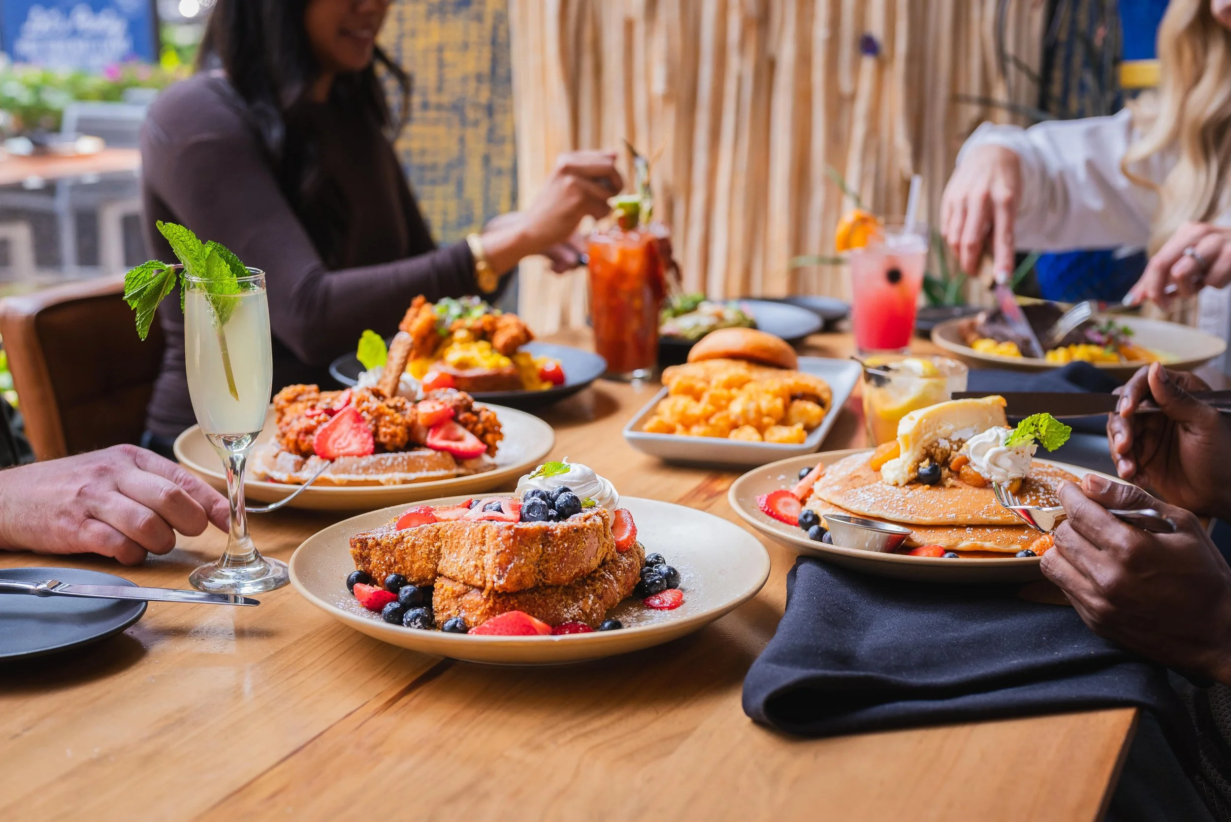 Group of friends enjoying brunch with plates of fried chicken, waffles, and colorful cocktails at a restaurant table.