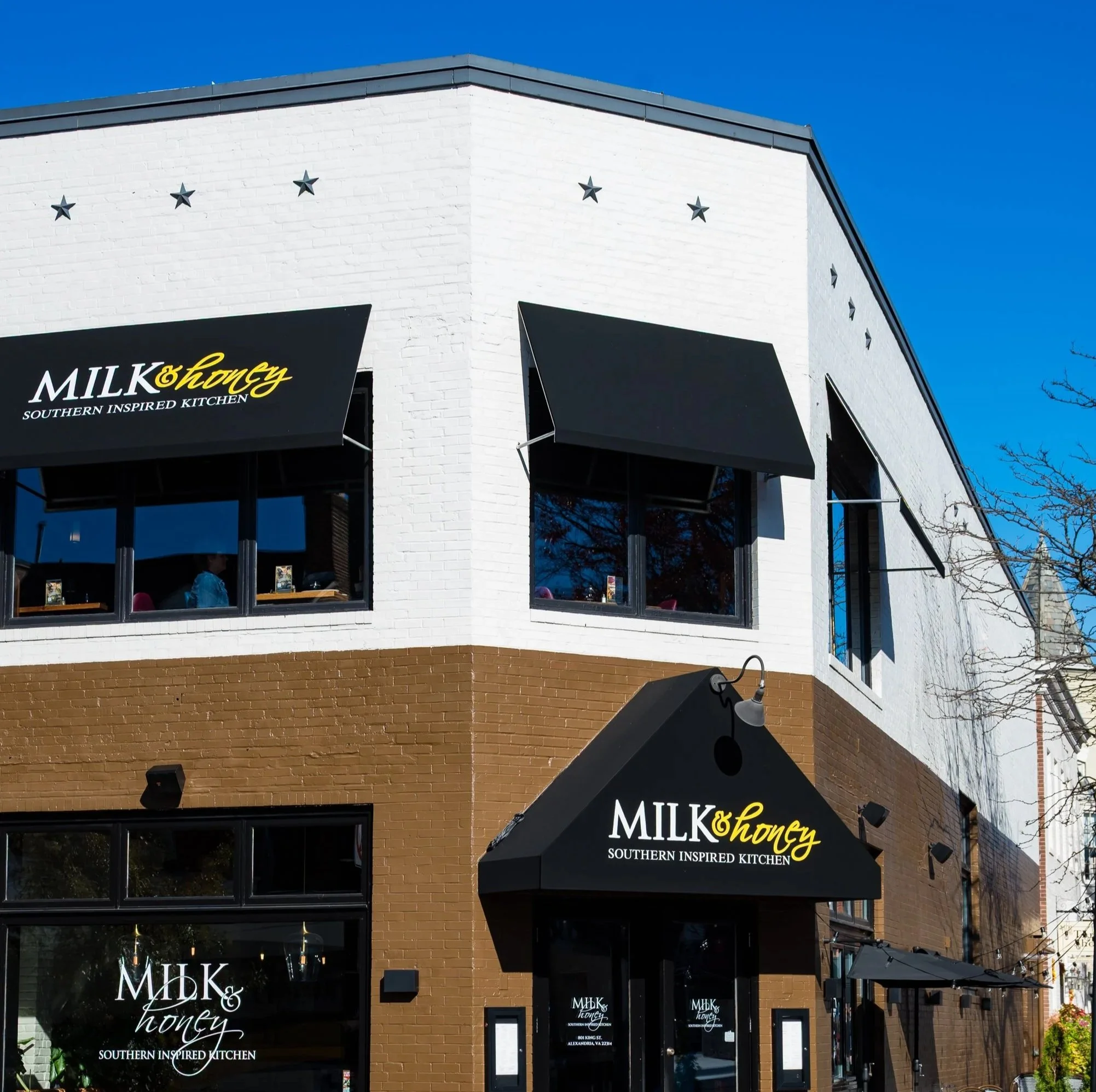 Corner view of Milk & Honey Southern Kitchen, a two-story building with white and brown brick exterior, black awnings, and outdoor seating on a sunny day.