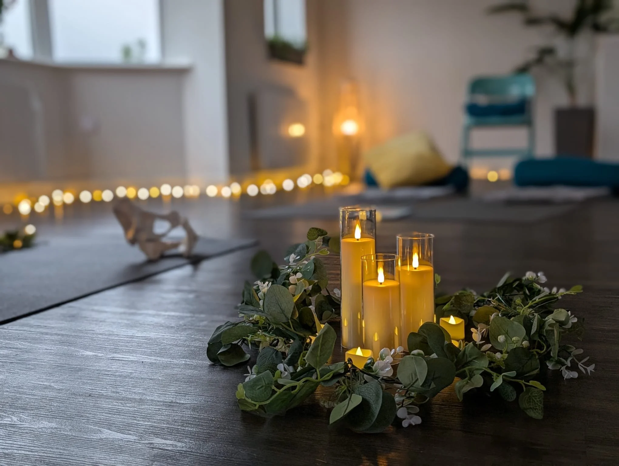 Decorative candles on a wooden floor surrounded by greenery and small white flowers in a cozy, softly lit room.