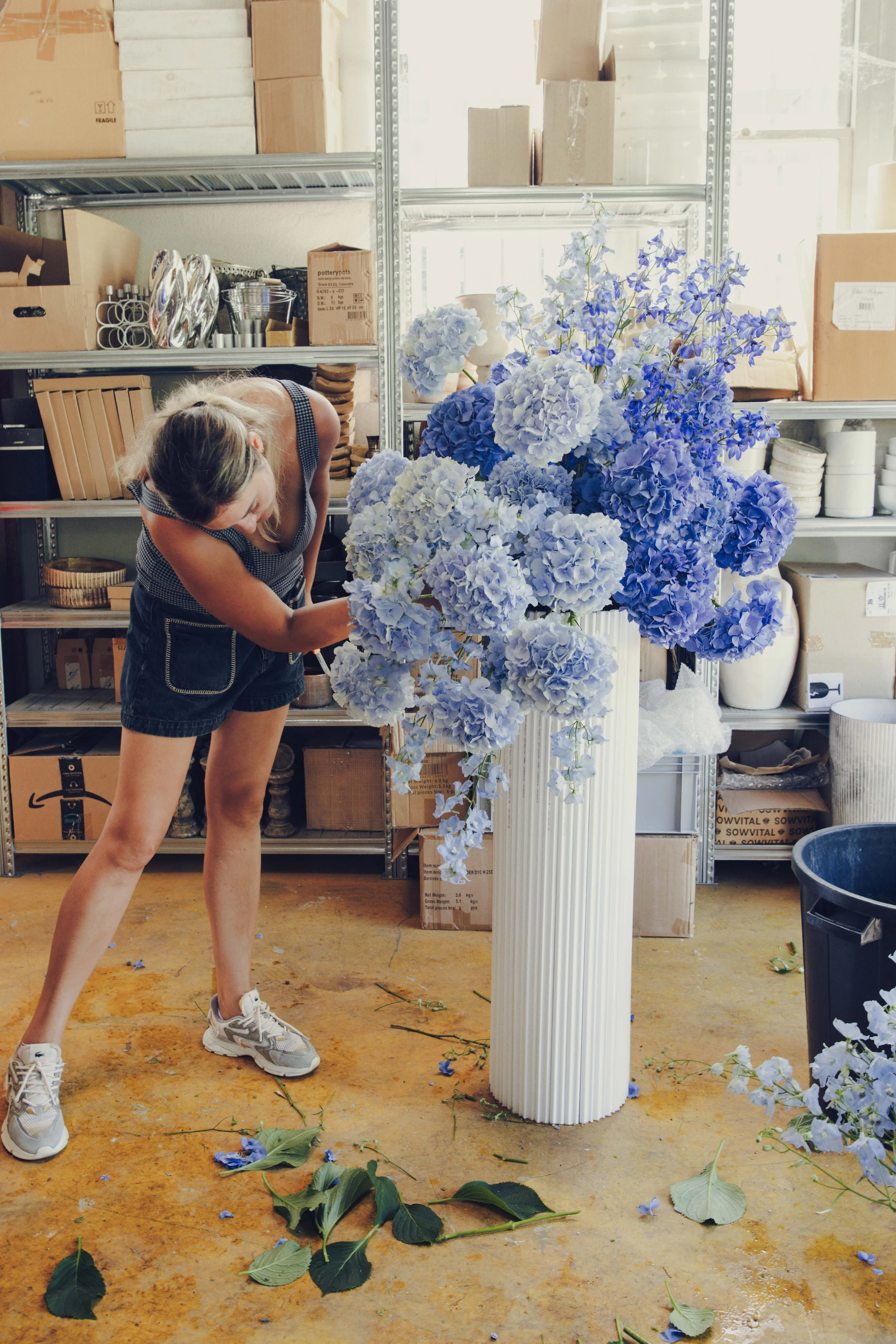 Florist arranging a tall blue hydrangea and delphinium design in a white fluted vase, event flowers studio, Côte d'Azur