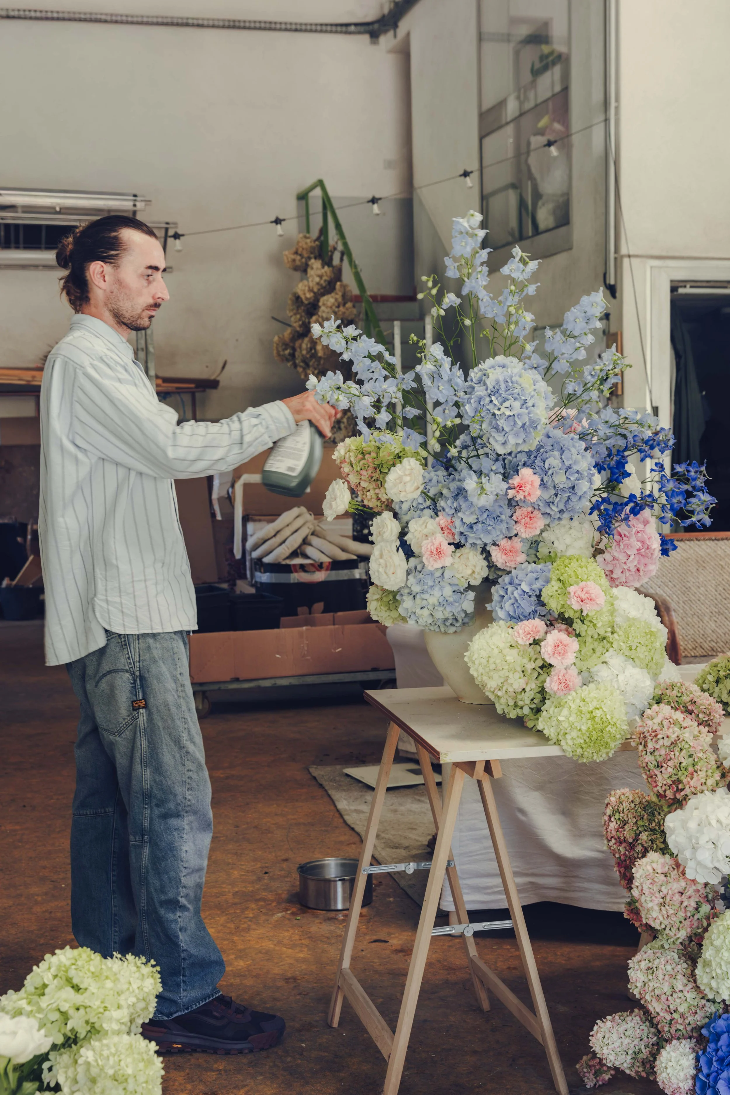 Florist tending a large pastel arrangement of blue hydrangea, pink roses and green hydrangea on a trestle table in the studio