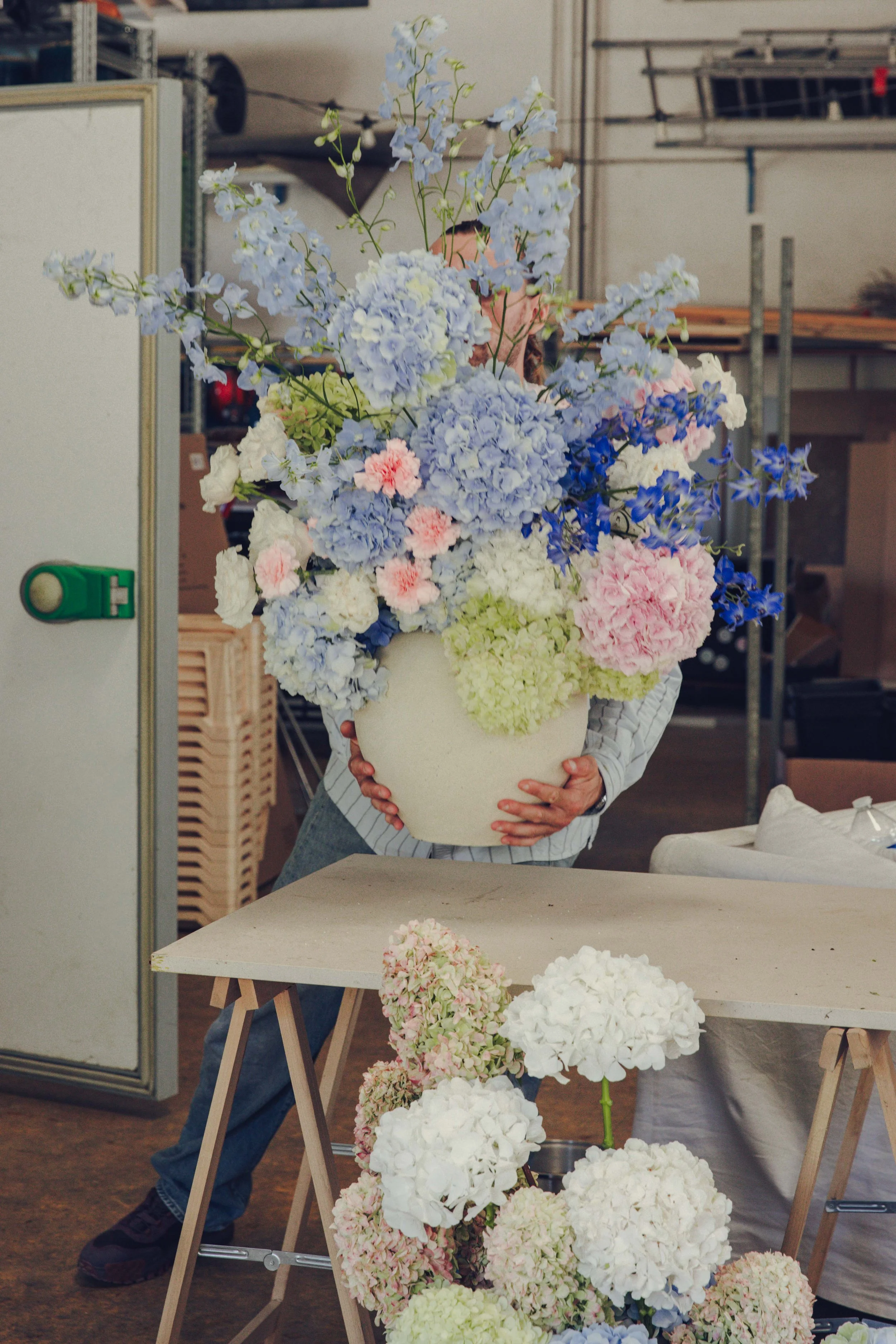 Florist holding a cream vase of blue, pink and green flowers, with white hydrangea on the studio worktable