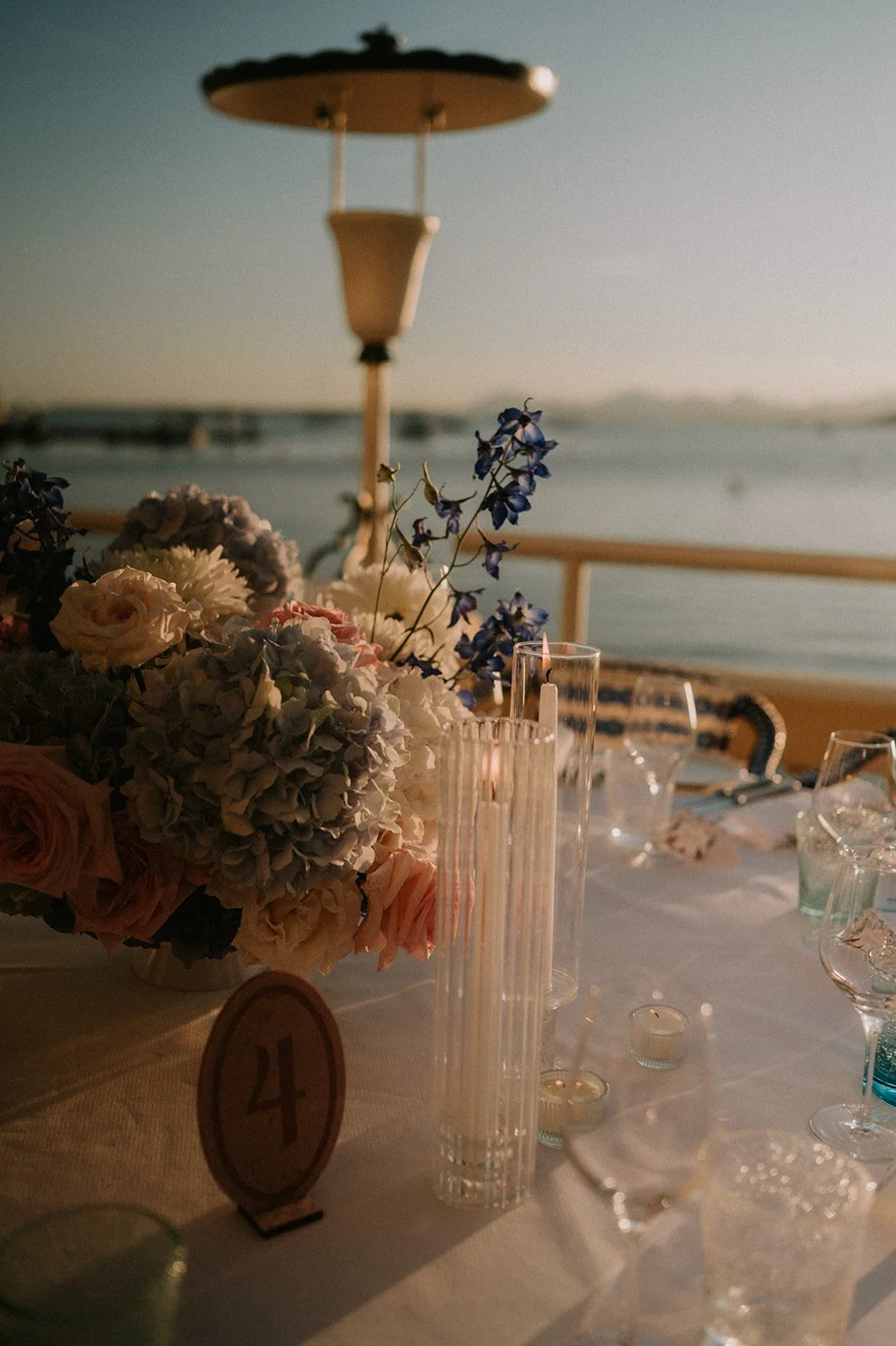 Wedding table number card beside a hydrangea, rose and delphinium centrepiece with fluted glass candle holders at sunset, Antibes