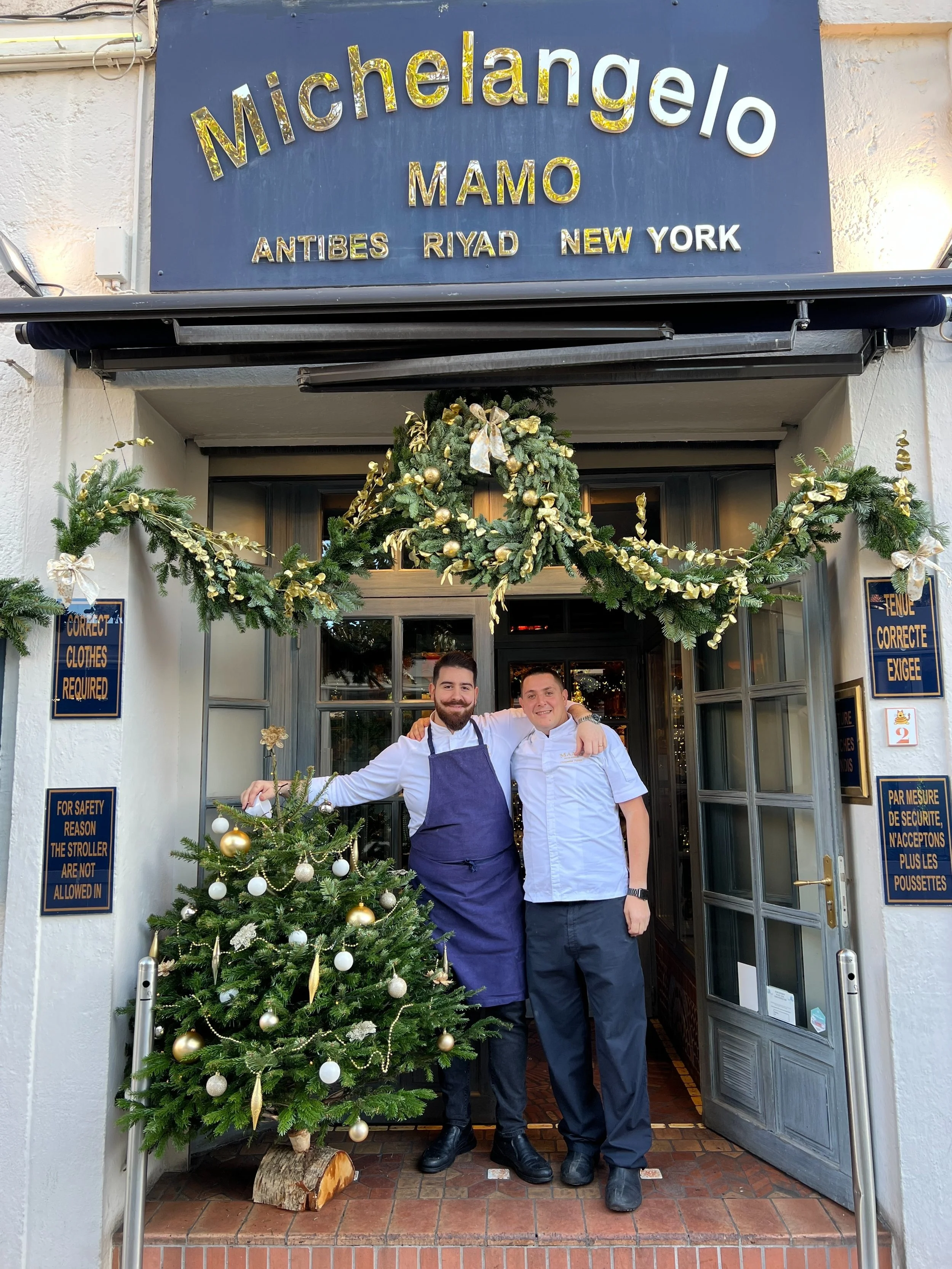 Restaurant entrance in Antibes decorated by GRÖNT with a fir and foliage garland and a Christmas tree with white ornaments, with two members of the restaurant team in the doorway