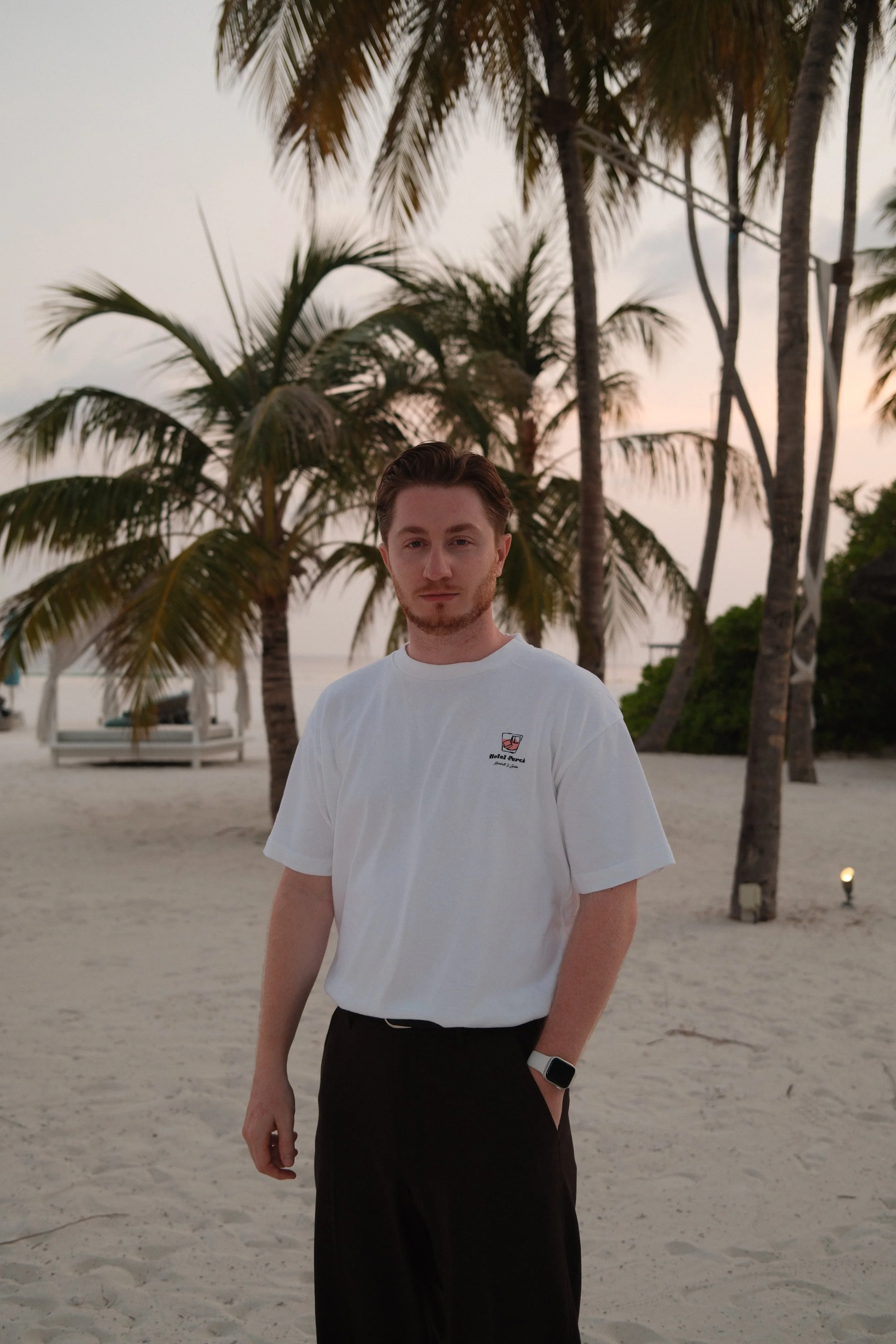 A young man with short brown hair and a light beard stands on a sandy beach with palm trees in the background, during sunset. He is wearing a white t-shirt and dark pants, with a smartwatch on his left wrist.