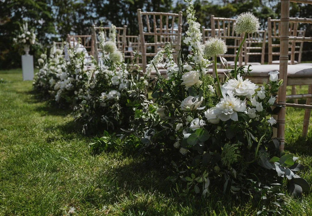 White flower arrangements with greenery along white chairs at an outdoor event, possibly a wedding, on a sunny day.