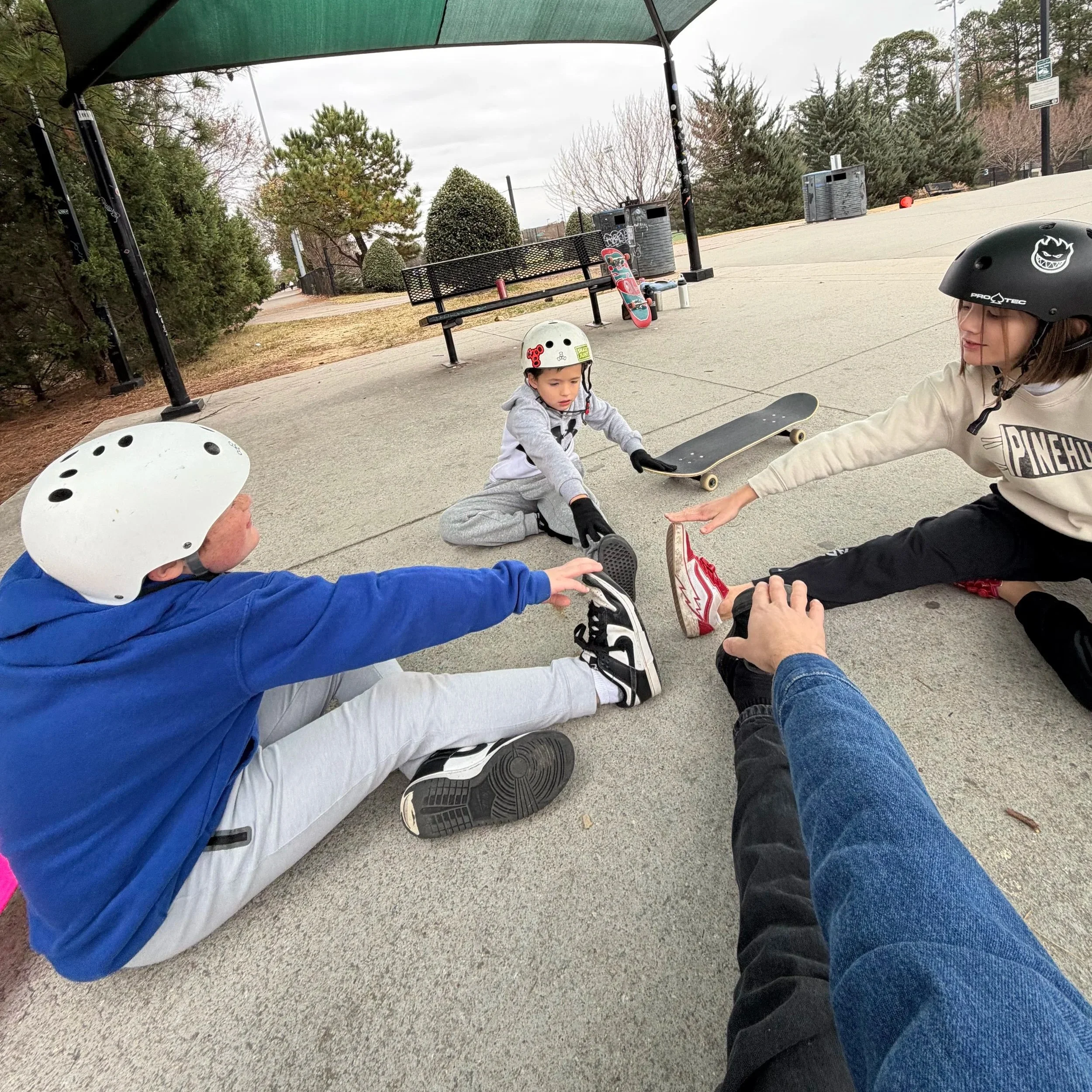 Four children wearing helmets stretching outdoors in a skate park under a canopy, with skateboards nearby, trees and a bench in the background.
