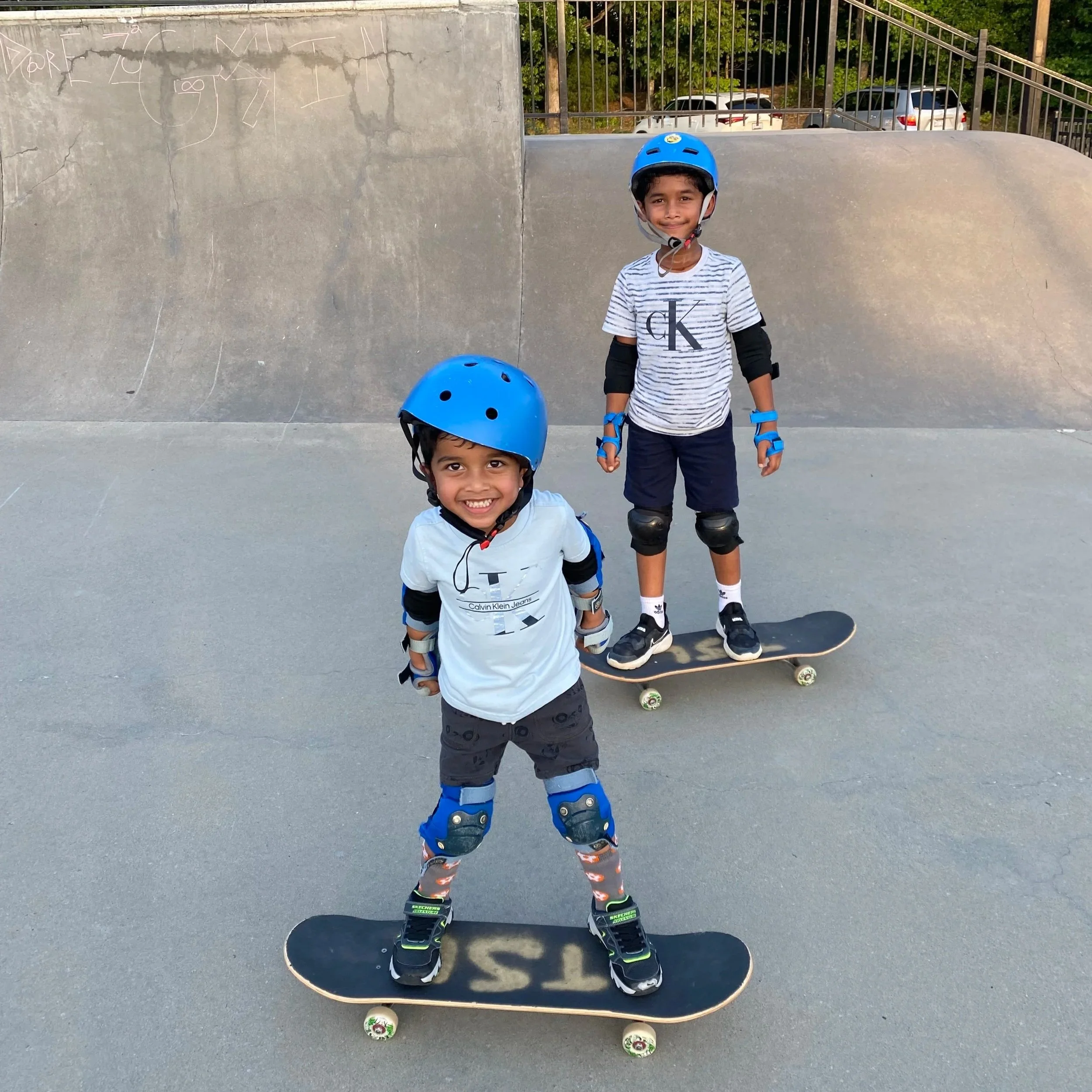 Two young boys wearing helmets and protective gear skateboarding at a skate park with a concrete ramp in the background.