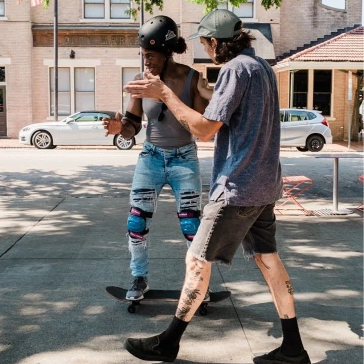 A woman with a helmet on a skateboard, wearing ripped jeans and knee pads, learns to skate with assistance from a man on a city sidewalk shade by trees, with buildings and parked cars in the background.