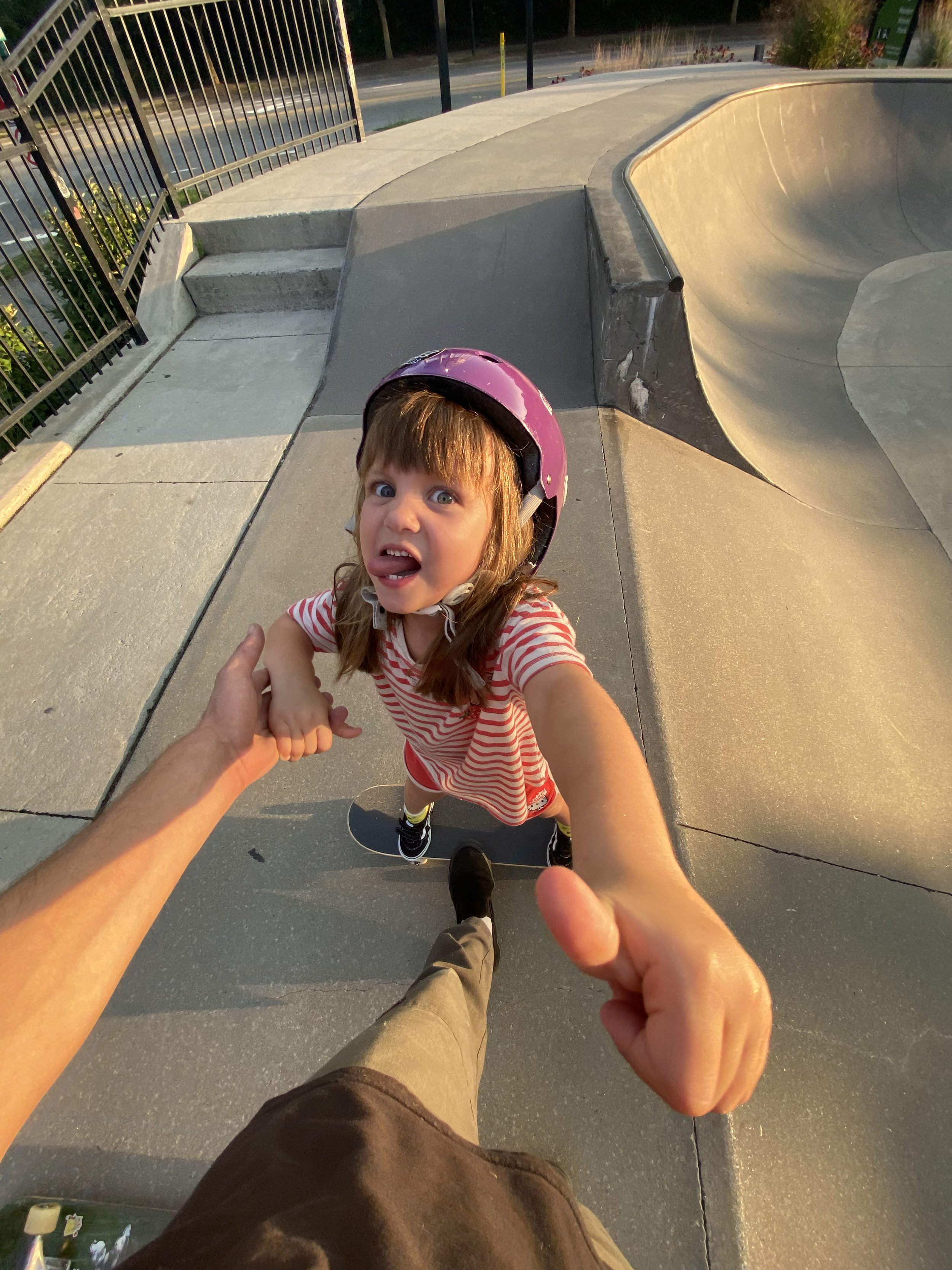 A young girl in a pink helmet making a funny face at a skate park, holding hands with an adult as she stands on a skateboard.