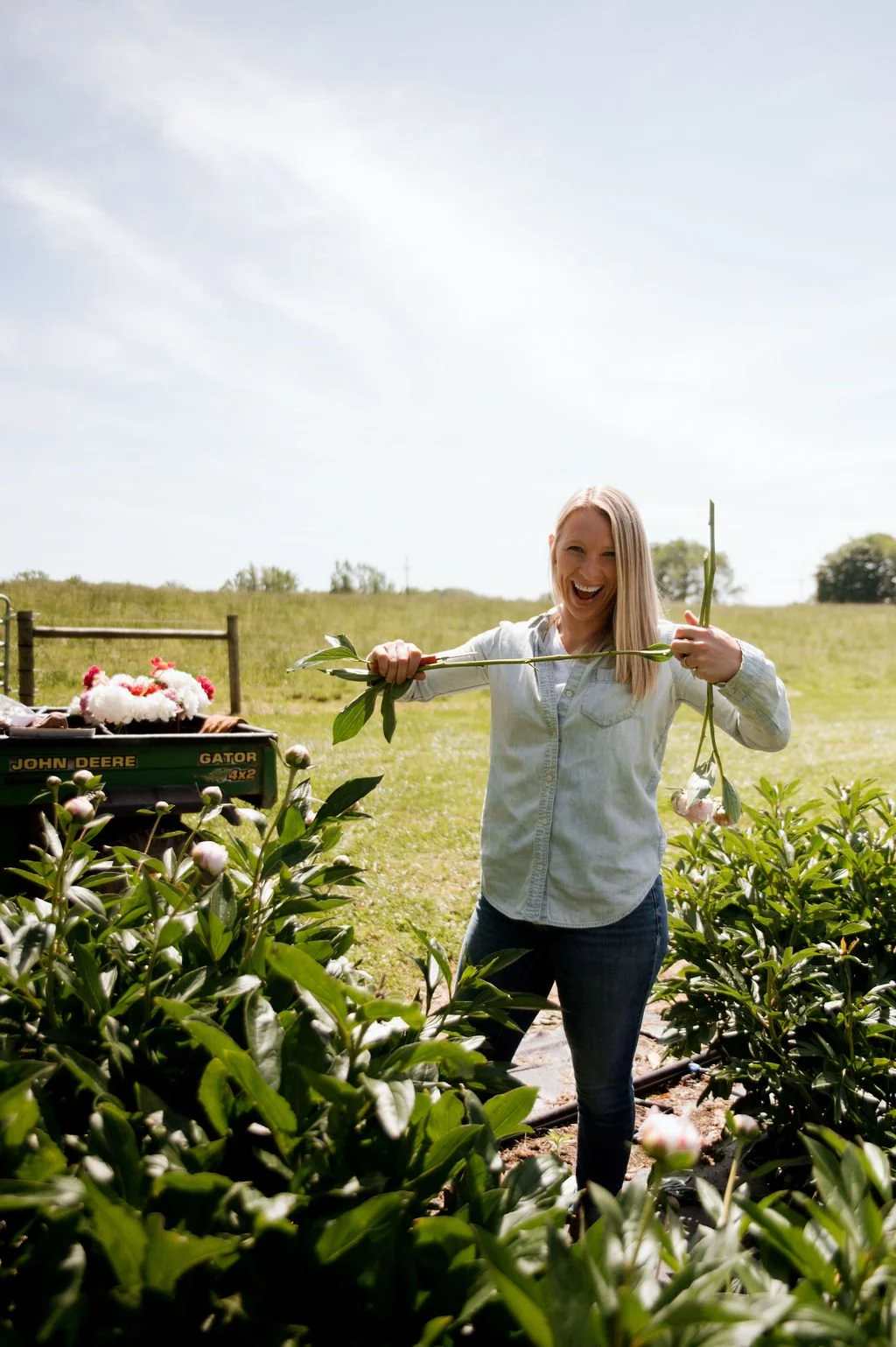 Jenny Marks harvesting peonies on her flower farm, smiling while cutting fresh stems in a peony field with a John Deere Gator in the background, showcasing real life flower farming work.