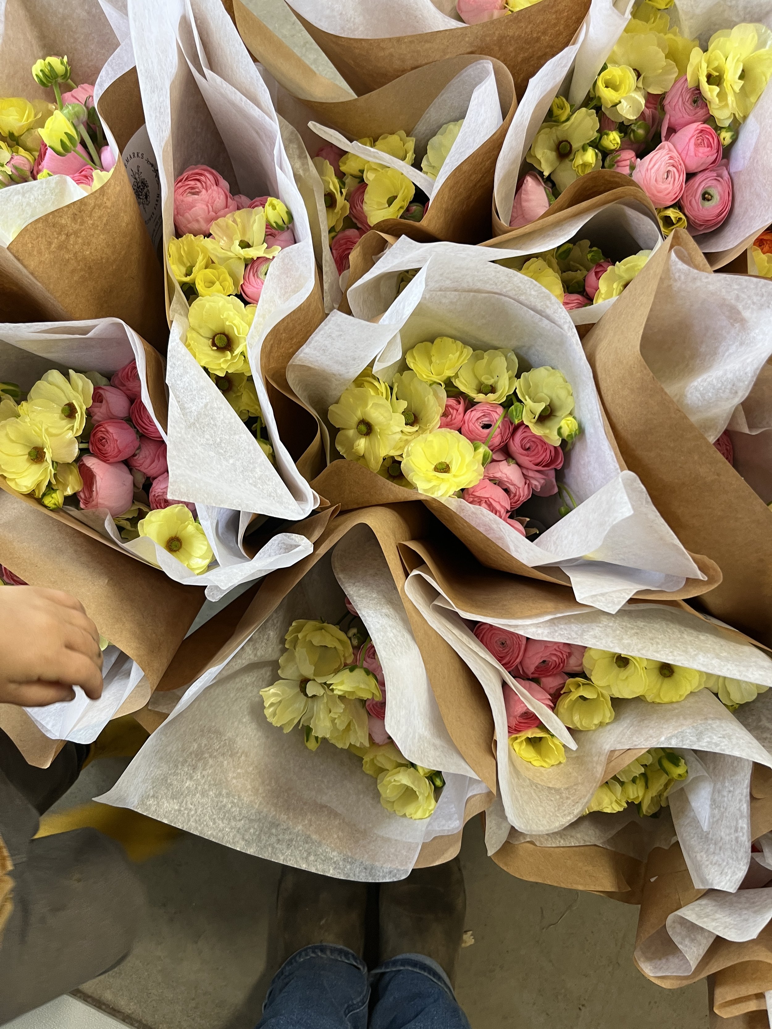 Jenny Marks of the Six Figure Flower Farming Podcast is pricing bouquets for profit on her flower farm. Yellow and pink ranunculus are wrapped with white and brown paper. The floor is abundant with cut flowers.